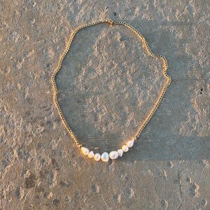 Puede incluir: Un collar de cadena dorada con una fila de perlas blancas. Las perlas son de diferentes tama&ntilde;os y est&aacute;n ensartadas a lo largo de la parte inferior de la cadena. El collar est&aacute; sobre una superficie clara y texturizada.