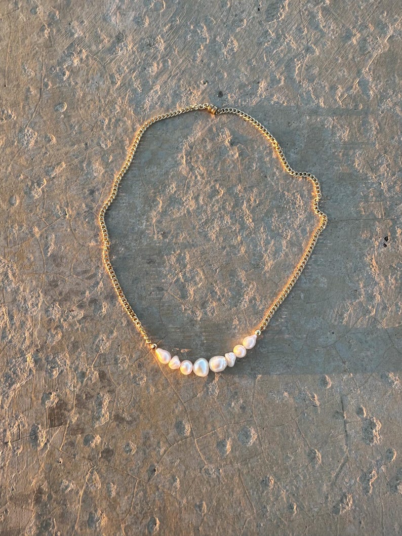 Puede incluir: Un collar de cadena dorada con una fila de perlas blancas. Las perlas son de diferentes tama&ntilde;os y est&aacute;n ensartadas a lo largo de la parte inferior de la cadena. El collar est&aacute; sobre una superficie clara y texturizada.