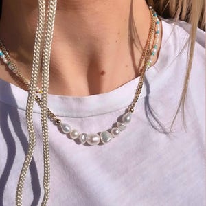 Puede incluir: Un collar de cadena dorada con una fila de perlas blancas irregulares. Tambi&eacute;n es visible un segundo collar de cadena dorada m&aacute;s fino con peque&ntilde;as cuentas turquesas y blancas. Un cord&oacute;n blanco cuelga en el lado izquierdo.