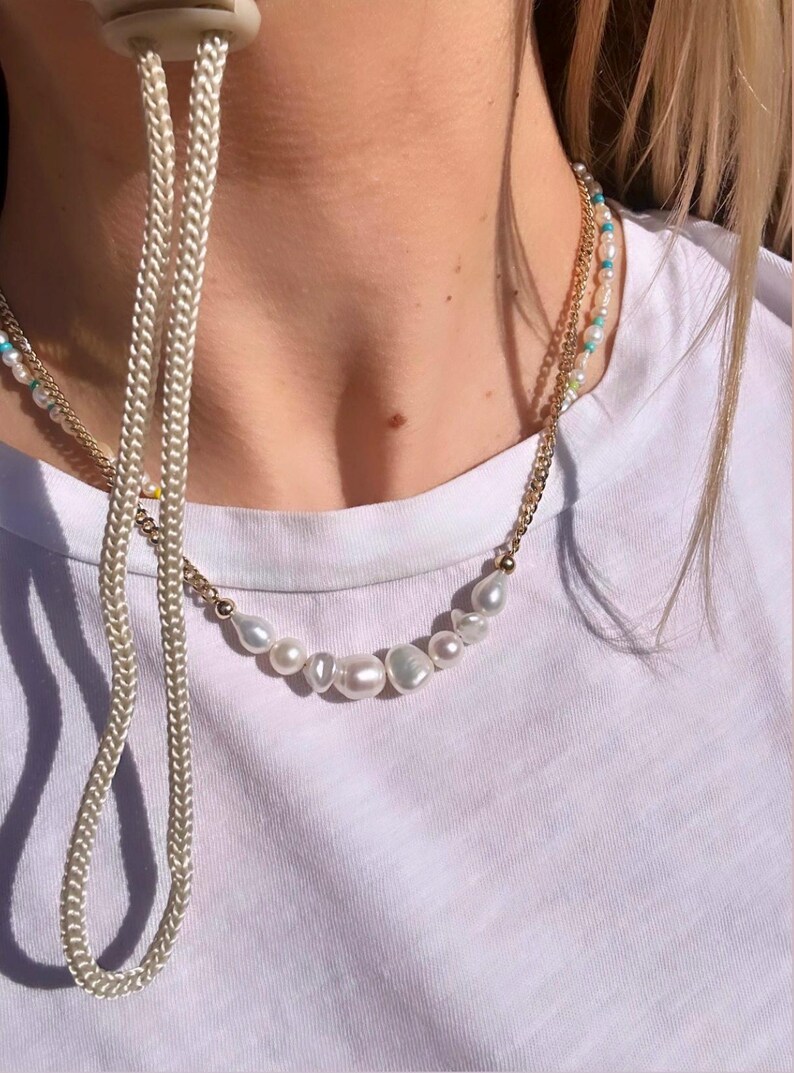 Puede incluir: Un collar de cadena dorada con una fila de perlas blancas irregulares. Tambi&eacute;n es visible un segundo collar de cadena dorada m&aacute;s fino con peque&ntilde;as cuentas turquesas y blancas. Un cord&oacute;n blanco cuelga en el lado izquierdo.