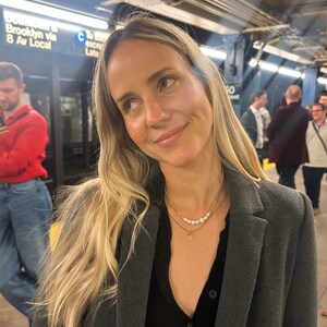 Puede incluir: Una mujer con cabello largo y rubio lleva un abrigo gris sobre una blusa negra. Lleva un collar dorado con perlas y un peque&ntilde;o colgante. El fondo muestra una estaci&oacute;n de metro con letreros.