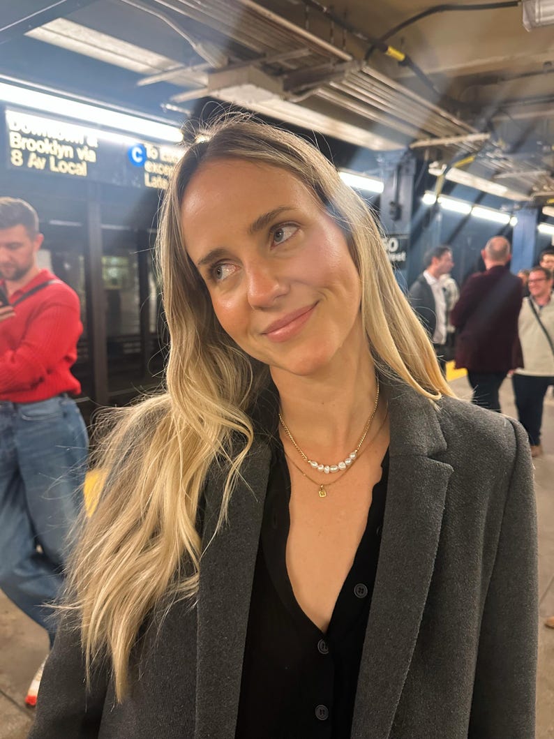 Puede incluir: Una mujer con cabello largo y rubio lleva un abrigo gris sobre una blusa negra. Lleva un collar dorado con perlas y un peque&ntilde;o colgante. El fondo muestra una estaci&oacute;n de metro con letreros.