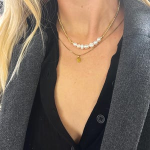 Puede incluir: Dos collares de oro. El collar superior presenta una hilera de perlas blancas. El segundo collar tiene un peque&ntilde;o colgante cuadrado de oro. Los collares est&aacute;n superpuestos a una camisa negra y un abrigo gris.