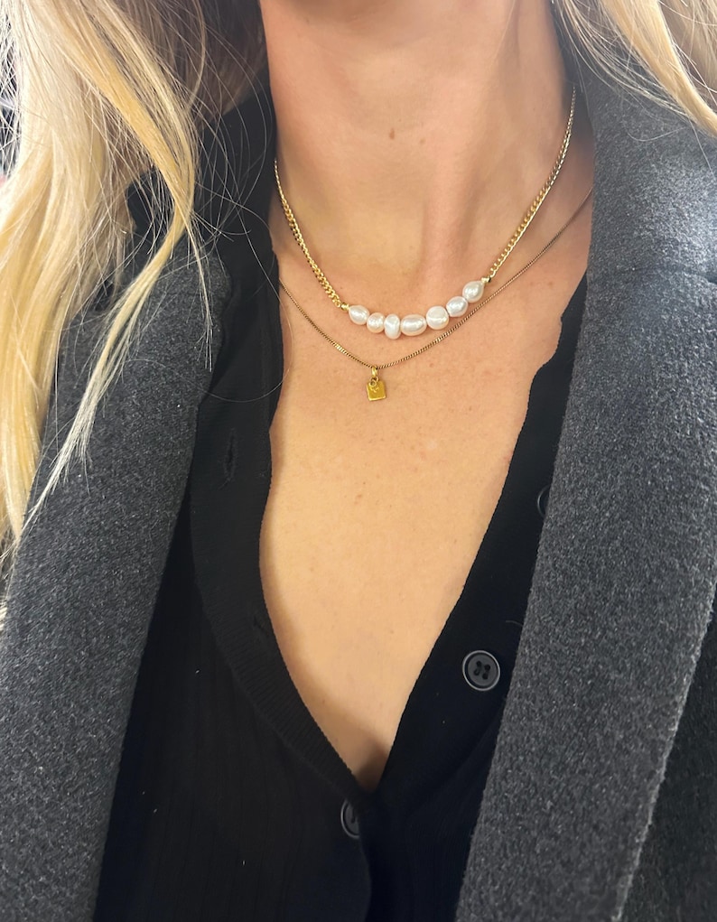 Puede incluir: Dos collares de oro. El collar superior presenta una hilera de perlas blancas. El segundo collar tiene un peque&ntilde;o colgante cuadrado de oro. Los collares est&aacute;n superpuestos a una camisa negra y un abrigo gris.