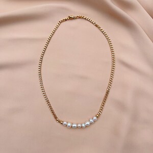 Puede incluir: Collar de cadena dorada con una fila de perlas blancas. El collar presenta un dise&ntilde;o de cadena de eslabones y un cierre de broche. Las perlas son de diferentes tama&ntilde;os y est&aacute;n ensartadas a lo largo de la parte inferior de la cadena.