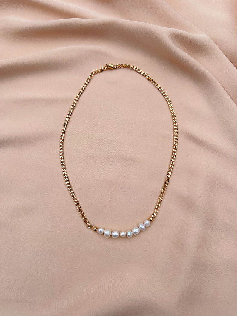 Puede incluir: Collar de cadena dorada con una fila de perlas blancas. El collar presenta un dise&ntilde;o de cadena de eslabones y un cierre de broche. Las perlas son de diferentes tama&ntilde;os y est&aacute;n ensartadas a lo largo de la parte inferior de la cadena.