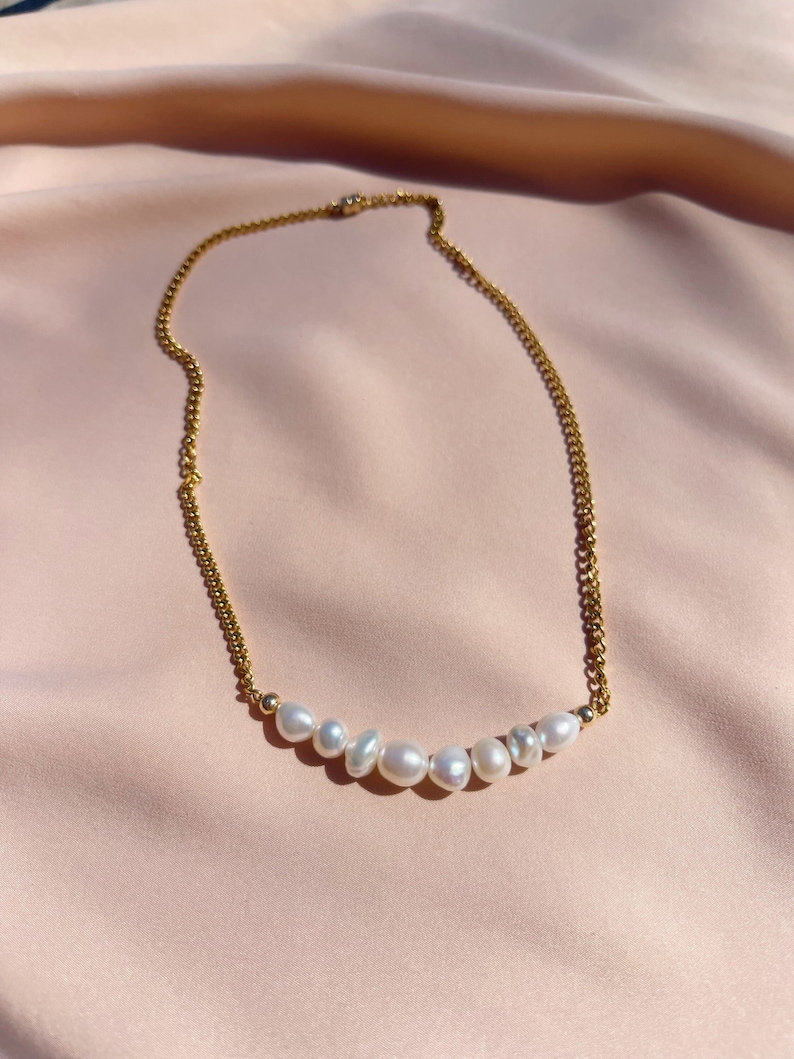 Puede incluir: Un collar de cadena dorada con una fila de perlas blancas. El collar se exhibe sobre una tela de sat&eacute;n rosa. Las perlas tienen formas irregulares y tama&ntilde;os variados, lo que a&ntilde;ade un toque &uacute;nico a la joya.