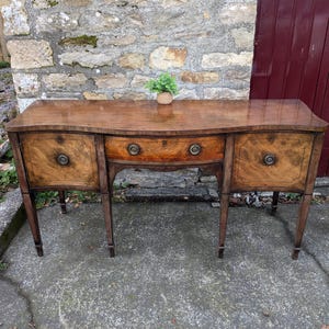 May include: A classic wooden sideboard with a curved front, featuring two side cabinets and a central drawer. The wood has a warm, reddish-brown tone with decorative grain patterns. The sideboard stands on slender, tapered legs and has brass hardware.