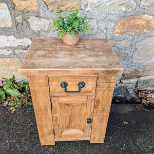 May include: A rustic wooden side table with a drawer and a small cabinet door. The table has a natural wood finish and a black metal handle. A small plant in a woven pot sits on top. The table is approximately 61 cm tall.