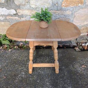 May include: An oval wooden table in light brown, with a small plant in a woven pot. The table has a four-legged base with turned legs and a distressed finish. The table is approximately 91 cm long.