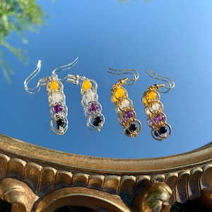 May include: Two pairs of drop earrings featuring a chainmaille design with yellow, white, purple, and black beads. One pair has silver-toned metal rings and hooks, while the other pair has gold-toned metal rings and hooks, both hanging from ear wires.