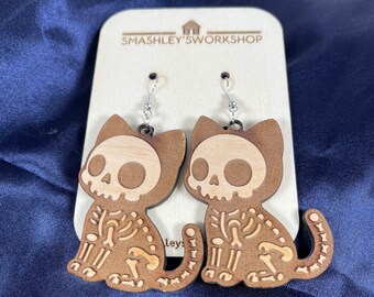 May include: A pair of wooden cat skeleton earrings with a brown wood finish. The earrings are shaped like a cat with a skull and bones design. The earrings are hanging from silver hooks. The earrings are labeled "Smashley's Workshop".