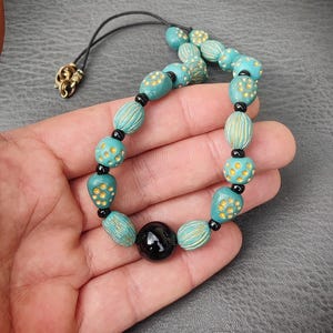 Può includere: Collana di perline turchesi e dorate con una perla di onice nera al centro. La collana è realizzata con perline in ceramica con motivi intricati.