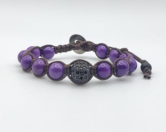 May include: A handcrafted bracelet with purple beads and a central black bead with a detailed pattern. The beads are strung on a brown cord, with a sliding knot closure. The bracelet is designed for a comfortable fit.