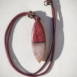 Può includere: Una collana con cordoncino in pelle marrone con un ciondolo in legno e resina. Il ciondolo ha la forma di una lacrima ed è composto da una parte superiore in legno scuro e una parte inferiore in resina trasparente con glitter argentati.