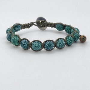 Puede incluir: Una pulsera de macramé con cuentas estampadas en verde y turquesa. Las cuentas están ensartadas en un cordón marrón, con una pequeña cuenta de madera y un cierre de botón plateado. La pulsera se muestra sobre una superficie blanca.