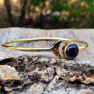May include: A gold bangle bracelet with a crescent moon charm and a round amethyst gemstone.