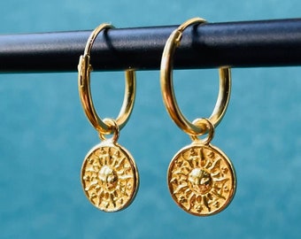 May include: A pair of gold hoop earrings with a sun charm pendant. The earrings are hanging from a black surface.