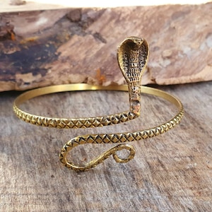 May include: A gold-toned metal bracelet shaped like a coiled snake. The bracelet features a textured, diamond-patterned body that wraps around the wrist, ending in a raised cobra head with detailed scales and a flared hood design.