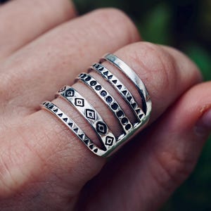 May include: A silver-toned multi-band ring featuring five distinct parallel bands connected at the base. Each band displays a unique engraved pattern, including triangles, circles, and diamond shapes, creating a layered appearance across the finger.