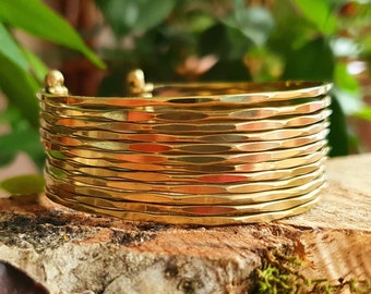 May include: A gold-colored cuff bracelet with a textured, hammered design. The bracelet is made of multiple stacked bands and has a simple, elegant style. The jewelry is displayed on a piece of wood with green foliage in the background.