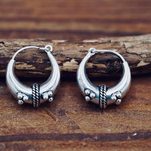 Op de afbeelding: Een paar zilverkleurige oorringen. Elke oorring heeft een halve maanvorm met een gevlochten band in het midden en drie kleine, ronde versieringen aan elke kant. De oorbellen worden gepresenteerd op een houten oppervlak.