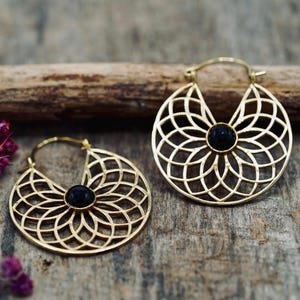 Puede incluir: Un par de pendientes de aro dorados con un diseño floral de filigrana y un centro de piedra negra. Los pendientes se exhiben sobre una superficie de madera con una rama seca y flores moradas.