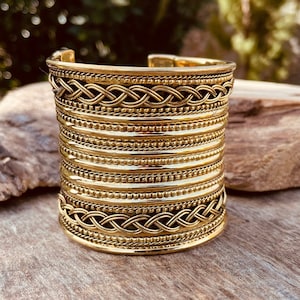 Puede incluir: Un brazalete ancho de color dorado con intrincados diseños trenzados y con cuentas. El brazalete presenta múltiples bandas horizontales y una superficie texturizada, creando una pieza audaz. El brazalete se exhibe sobre una superficie de madera.