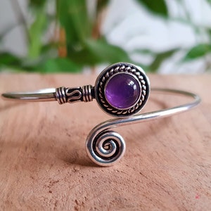 May include: A silver arm cuff bracelet with a central, round amethyst gemstone. The bracelet has a spiral design at one end and decorative detailing near the gemstone setting. The amethyst is a deep purple colour.