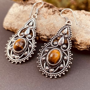 Può includere: Un paio di orecchini pendenti in argento con una pietra di occhio di tigre in ogni orecchino. Gli orecchini hanno un design delicato e ornato.