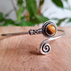 Puede incluir: Un brazalete de plata con un diseño de espiral y una piedra redonda de ojo de tigre.