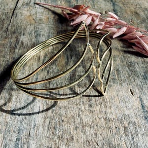 Puede incluir: Un brazalete tipo brazalete de metal en tono dorado con un diseño de múltiples capas de bandas de alambre finas y curvas que forman una forma geométrica abierta. El brazalete tiene un acabado pulido y una estructura flexible y abierta.