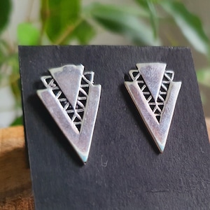 Op de afbeelding: Een paar zilveren oorbellen met een geometrisch driehoeksontwerp. De oorbellen worden getoond op een zwarte achtergrond.