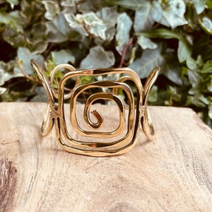 Può includere: Un bracciale a fascia in metallo color oro caratterizzato da un design centrale a spirale quadrata. Il bracciale presenta una superficie lucida e riflettente e una struttura aperta e curva che si estende da entrambi i lati dell'elemento.