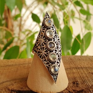 Könnte beinhalten: Ein silberner Ring mit geometrischem Design und fünf Mondstein-Steinen. Der Ring wird auf einem hölzernen Ringständer präsentiert.
