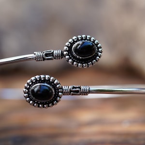 May include: A silver open cuff bracelet featuring two oval black stones, each set in a beaded silver frame. The band has a textured, wire-wrapped detail near the stone settings, creating a symmetrical design with polished silver metal ends.