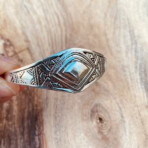 May include: A silver bracelet with a geometric design. The bracelet is made of metal and has a diamond-shaped center piece.