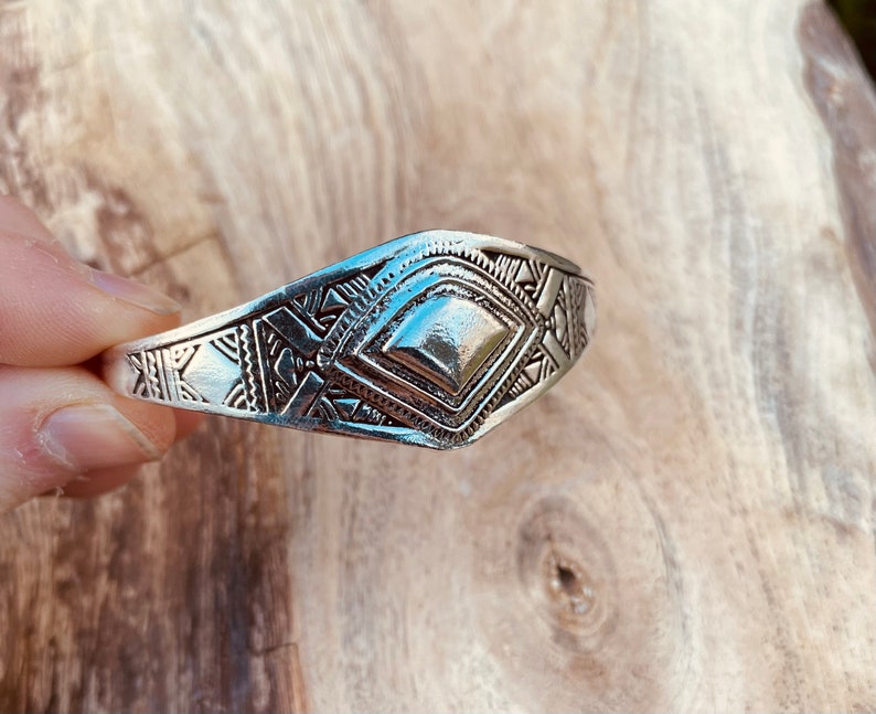 May include: A silver bracelet with a geometric design. The bracelet is made of metal and has a diamond-shaped center piece.