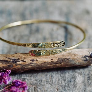 Peut inclure: Un bracelet manchette en métal doré composé d'un anneau fin et circulaire se terminant par deux extrémités aplaties qui se chevauchent. La surface de ces deux extrémités présente une finition martelée et texturée qui capte la lumière.