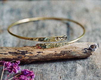 May include: A gold-toned metal cuff bracelet featuring a thin, circular band that transitions into two flattened, overlapping ends. The surface of these two ends displays a distinct, hammered, and textured finish that catches the light.