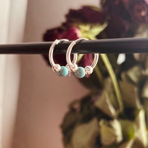 May include: A pair of silver hoop earrings with turquoise beads. The earrings are hanging from a black rod.