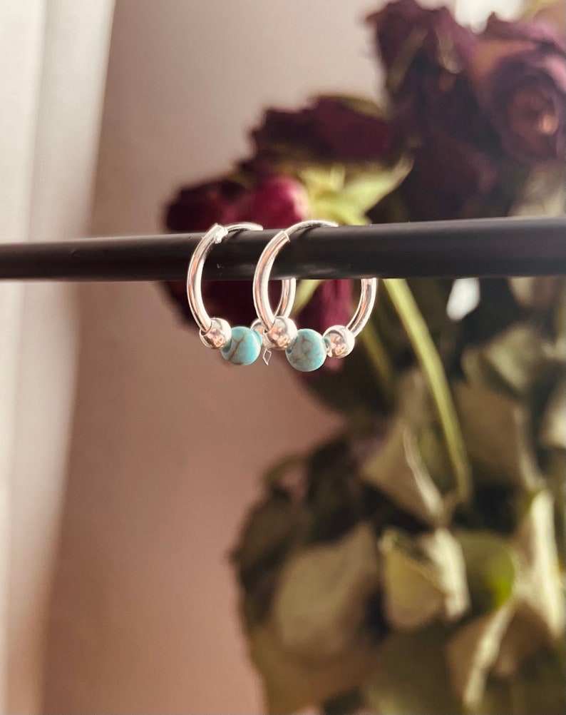 May include: A pair of silver hoop earrings with turquoise beads. The earrings are hanging from a black rod.