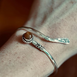 Op de afbeelding: Een zilverkleurige open armband met een gepolijste, ronde, barnsteenkleurige edelsteen in een kraalvormige zetting. De metalen band heeft gedetailleerde draadwikkelingen bij de steen en een getextureerde, gehamerde afwerking aan het uiteinde.