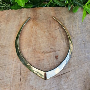 May include: A silver choker necklace with a V-shaped design. The necklace is made of a smooth, shiny metal and is lying on a wooden surface.