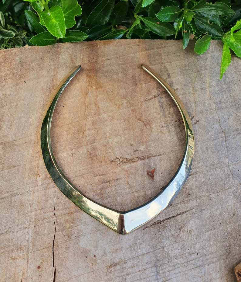 May include: A silver choker necklace with a V-shaped design. The necklace is made of a smooth, shiny metal and is lying on a wooden surface.