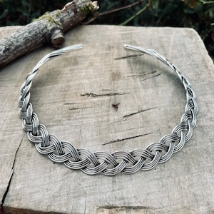 Puede incluir: Un collar de plata trenzado con un diseño sencillo. El collar está hecho de un alambre trenzado grueso y tiene un acabado liso y pulido.