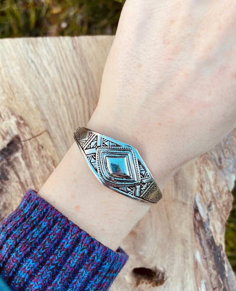 May include: A silver bracelet with a geometric design. The bracelet is made of metal and has a diamond-shaped center piece.