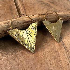 May include: A pair of gold-colored, triangular earrings with intricate etched patterns. The earrings feature a geometric design with a series of small circles along the edges and a sunburst pattern in the center. The earrings are displayed on a wooden surface.