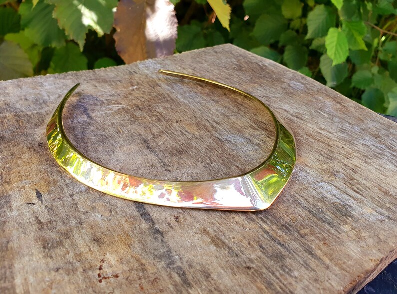 May include: A gold-toned metal choker necklace with a wide, flat, and slightly pointed design. The necklace is made of a hammered metal, giving it a textured surface.