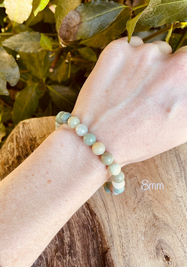 May include: A bracelet made of light green, white, and beige round beads. The beads are approximately 8mm in diameter. The bracelet is worn on a person's wrist.